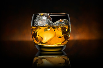 Single glass of whisky whiskey bourbon on ice with a reflective black surface and wooden background