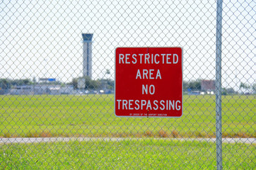 A RESTRICTED AREA NO TRESPASSING sign on a fence at an airport with the air traffic control tower in the background