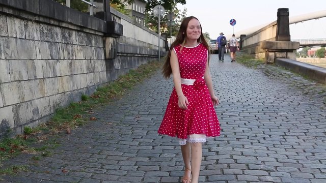 Young pretty girl in a red dress goes merrily skipping along pavement city waterfront