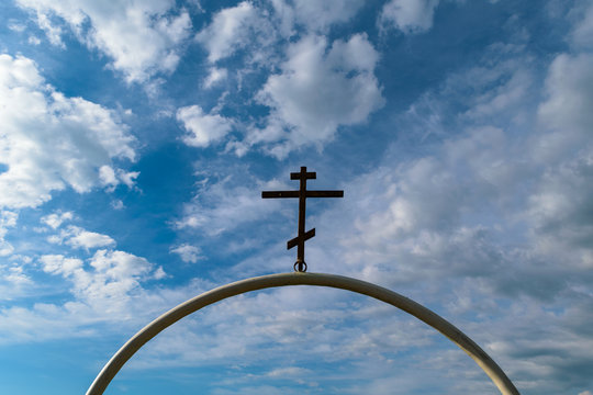 White semi-circular arch of iron pipe with dark Orthodox cross on it against the blue sky with clouds