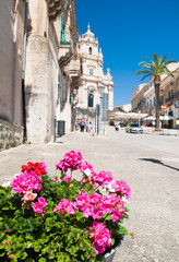 Rustic metal vase with pink geraniums and the baroque Saint George church in the background, Ragusa Ibla, East Sicily