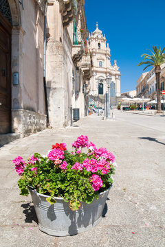 Rustic metal vase with pink geraniums and the baroque Saint George church in the background, Ragusa Ibla, East Sicily