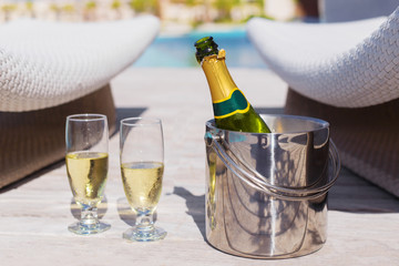 Cold champagne bottle in ice bucket and two glasses of champagne on the deck by the swimming pool
