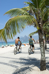 Pair of Brazilians riding bicycles along the boardwalk at Arpoador along a view of Ipanema Beach in Rio de Janeiro, Brazil