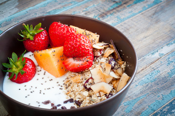 A bowl of healthy breakfast muesli with yogurt and topped with fresh strawberries, sliced melon, goji berries, carob and coconut shavings.