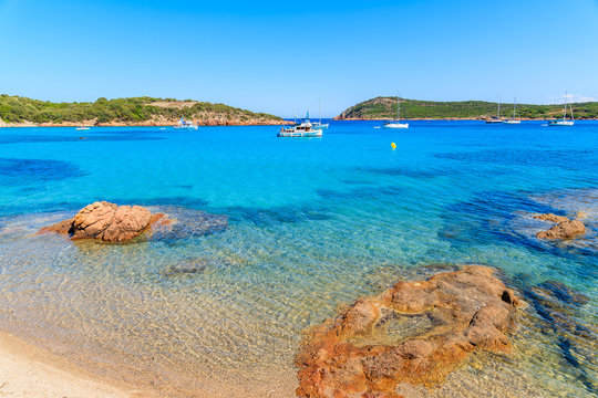 Red color rocks on Santa Giulia beach with boats on azure sea water, Corsica island, France