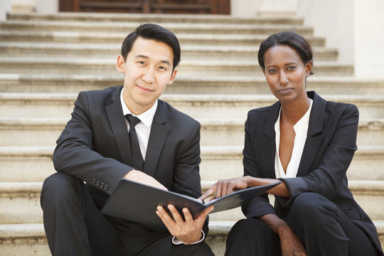 Professionally Dressed Man And Woman Sitting On An Outdoor Staircase Make Eye Contact With The Viewer.