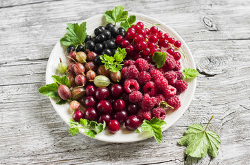 berries - raspberries, gooseberries, red currants, cherries, black currants on a white plate on a light wooden background