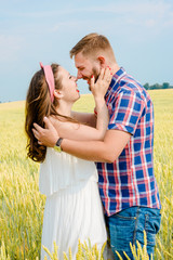 Happy young adult couple in love spending time on the wheat field on sunny day outdoors. Handsome man and beautiful woman standing and kissing. Romance and love concept
