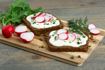 Two sandwiches with sour cream, onions and radishes and dill on a cutting board on a brown wooden...