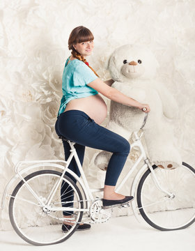 Toned photo of pregnant woman riding on bike with big teddy bear