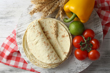 Stack of homemade whole wheat flour tortilla and vegetables on cutting board, on wooden table background