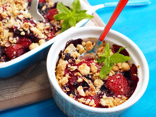 Crumble with strawberries, red currants, blueberries and blackberries with crumb made of powdered sugar, almonds, flour and oatmeal flakes in a ovenproof bowl with a spoon inside