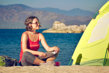 Young lady hiker sit near the tent and looking on sea landscape.