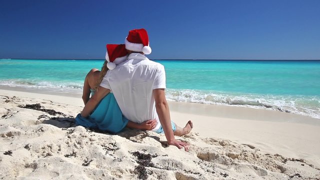 Couple in Santa Claus hats sitting on tropical sandy beach with turquoise sea water
