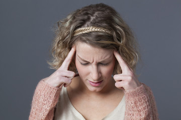 frowning young girl with curly blonde hair and pink sweater looking down with hands on temples for seeking for concentration or fighting against migraine