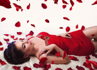 Obraz na płótnie Canvas Sexy beautiful brunette woman in red dress laying on a bed under romantic falling rose petals 