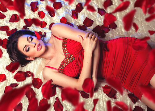 Sexy beautiful brunette woman in red dress laying on a bed under romantic falling rose petals 
