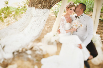 Fototapeta na wymiar beautiful gorgeous blonde bride and stylish groom on the decorated swing on the tree, wedding ceremony on cyprus