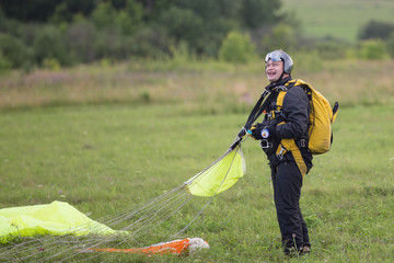 Joyful man in black suit with yellow parachute standing on green field after jump