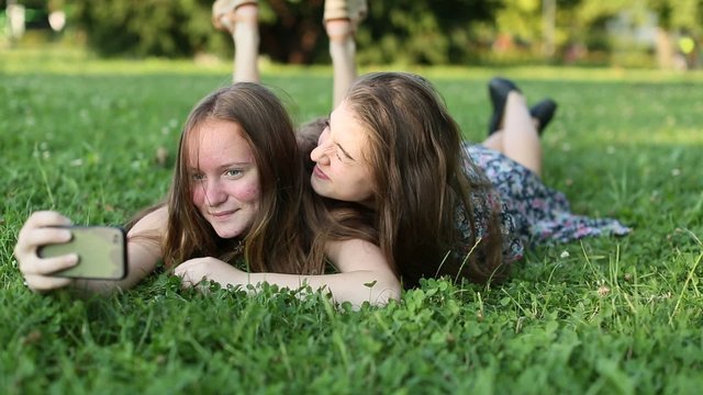 Two young girls best friends lying on the green grass and fun take a selfie on smartphone.