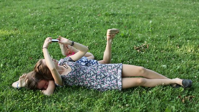 Two young girls best friends lying on the green grass and fun take a selfie on smartphone.