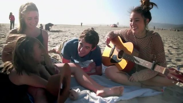 Group of teenage friends sit on the beach together listening to the guitar and messing around with each other  