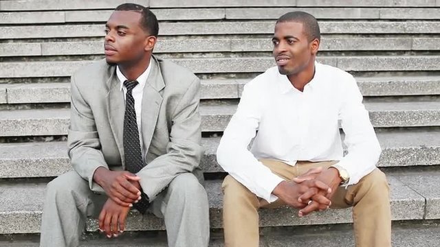 Two handsome black professionals sit on granite steps and one points as they listen to a street performer.