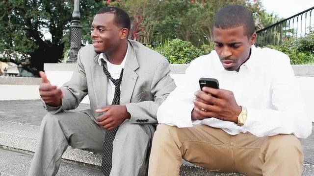 Two black male professionals listen to a street performer while one friend texts on his phone