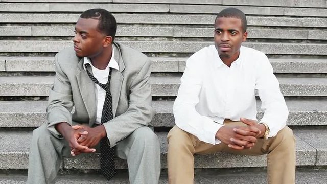 Two black professionals nod their heads and listen to a street performer while they sit on steps wearing business clothes
