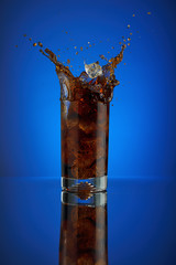 Splash of cola soft drink on a blue background. Refreshing  liquid drink coca pouring into a glass with ice. Pour high speed beverage for promoting restaurant and bar. isolated  liquor.
