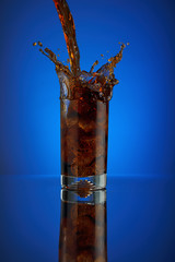 Splash of cola soft drink on a blue background. Refreshing  liquid drink coca pouring into a glass with ice. Pour high speed beverage for promoting restaurant and bar. isolated design
