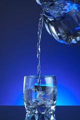 Pitcher water poured into a glass, splash, blue background, refreshing, freshness and health. Water bottle, water pitcher, blue liquid, ice, drops, motion, wave, splash, transparent blue water,
