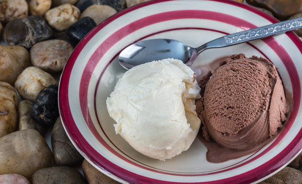 horizontal image of a white bowl with pink rim with two scoops of chocolate and a vanilla ice cream balls lying next to each other with a spoon sitting on a bed of pebbles.