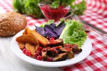 Beef with cranberry sauce, roasted potato slices, vegetables and bun on plate, on color wooden background