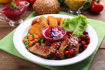 Beef with cranberry sauce, roasted potato slices, vegetables on plate, on  wooden background