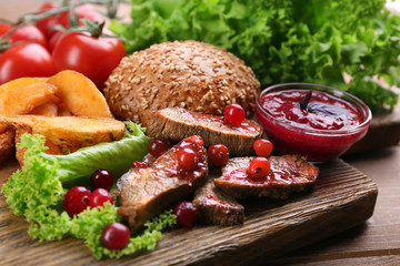 Beef with cranberry sauce, roasted potato slices and bun on cutting board, on wooden background