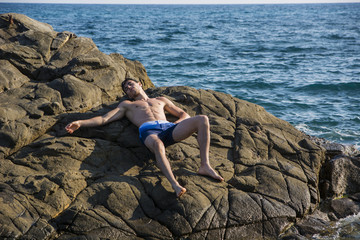Young shirtless athletic man leaning on rock by water on ocean