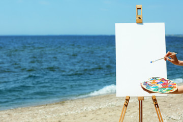 Female hand holding palette with paints and easel with canvas on beach