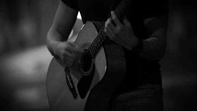 Black and white: long-haired (with a "beatle-like" hairdo) teenager performing an acoustic song playing the guitar on an empty moonlit street.