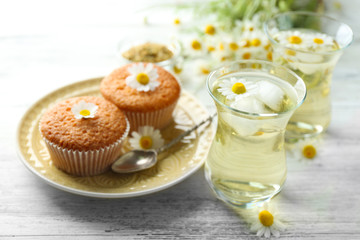 Glasses of chamomile tea with chamomile flowers and tasty muffins on color wooden background