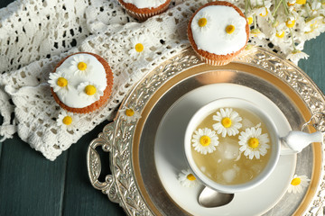 Cup of chamomile tea with chamomile flowers and tasty muffins on tray, on color wooden background