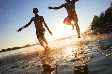 Glückliche junge Menschen laufen und springen am See beim Sonnenuntergang - obrazy, fototapety, plakaty