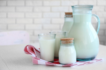 Pitcher, jars and glass of milk on wooden table, on bricks wall background