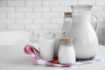 Pitcher, jars and glass of milk on wooden table, on bricks wall background