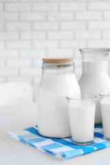 Pitcher, jar and glass of milk on wooden table, on bricks wall background