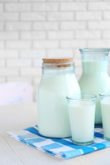 Pitcher, jar and glass of milk on wooden table, on bricks wall background