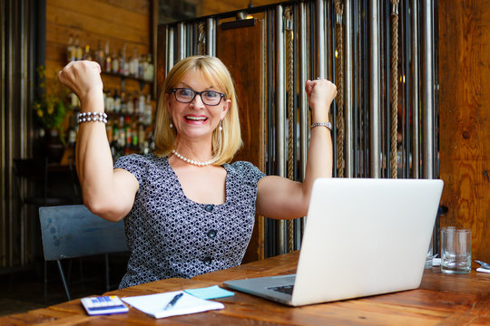 Close Up Portrait Smiling Old Senior Blonde Business Woman In The Glasses, Alone With Laptop In Summer Cafe With Gesture On Face And Raised Hands Up With Fists