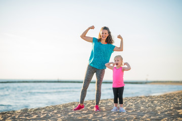 Happy mother and daughter in fitness gear on beach flexing arms