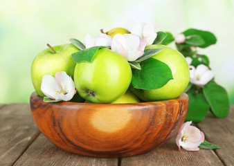 Fresh apples with apple blossom in bowl, on wooden table, on nature background